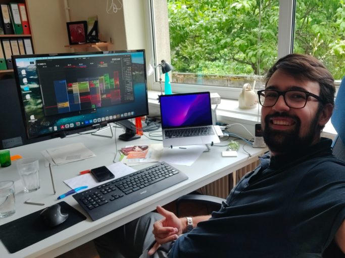 Im JPZ Felix sitzt lächeln an einem Schreibtisch mit zwei Bildschirmen und Pflanzen im Hintergrund.