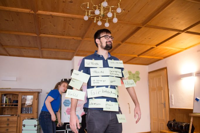 JuLeiCa-Einheit - Der*die ideale Leiter*in Mann mit Zetteln am Körper steht im Raum, während eine Frau im Hintergrund agiert.