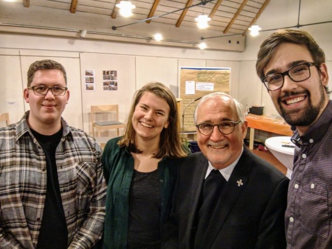 "Wie siehts aus Herr Bischof?" Mit Bischof Fürst Drei Personen lächeln zusammen mit einem Bischof in einem Innenraum, mit Tischen und Stühlen im Hintergrund.