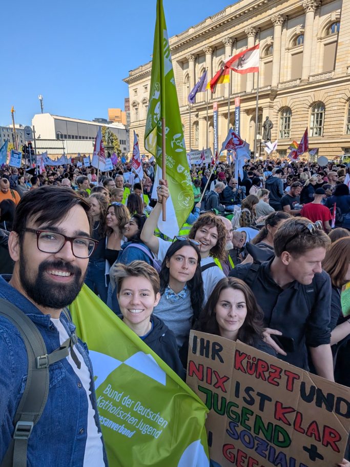 Auf Demo Felix posiert vor einer Menschenmenge mit Protestschildern und Fahnen.