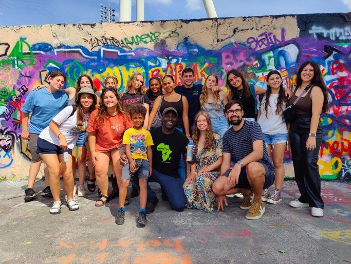 Meine erste IB - I feel you 2023 Gruppenfoto von Menschen vor einer bunten Graffiti-Wand, alle lächeln.